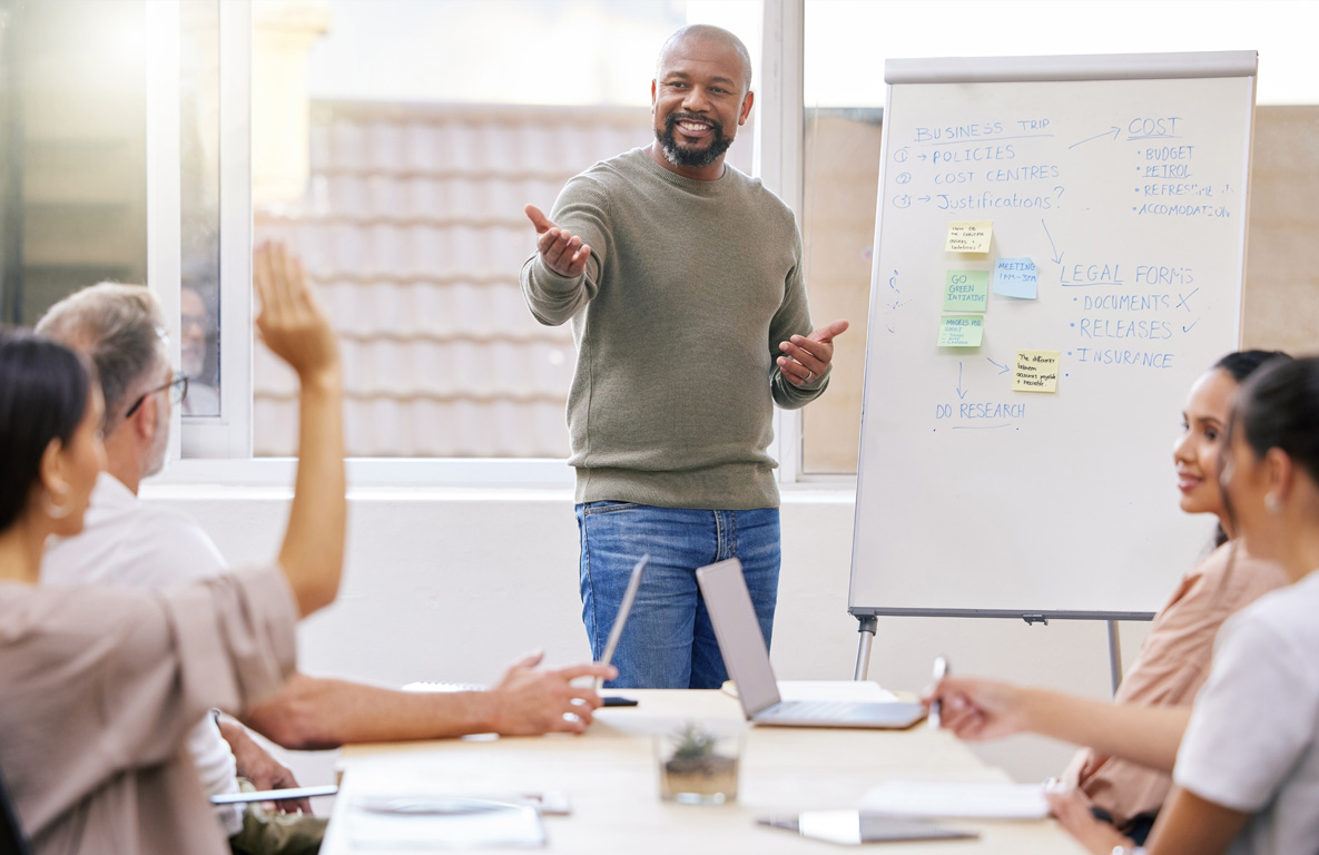 a man standing next to a white board in front of a conference table of people