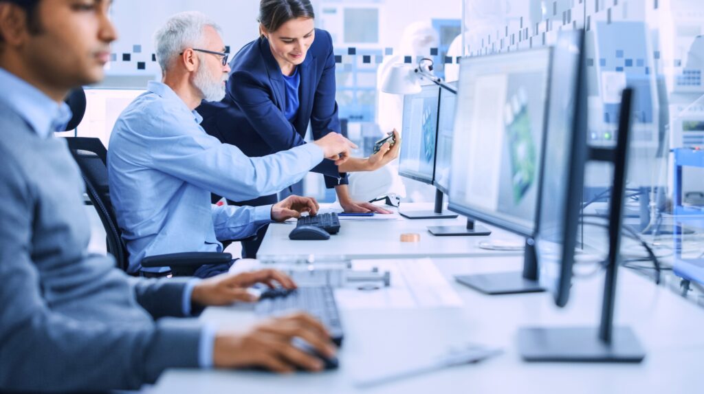 Female employee talks to senior male who is sitting at his desk, another employee is in the foreground