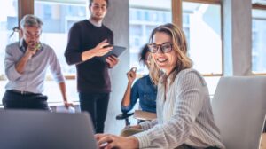 smiling person working on laptop in office while coworkers look at computer behind her