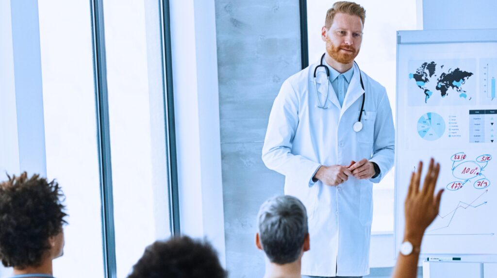 Doctor is presenting in front of other medical professionals and one has their hand up