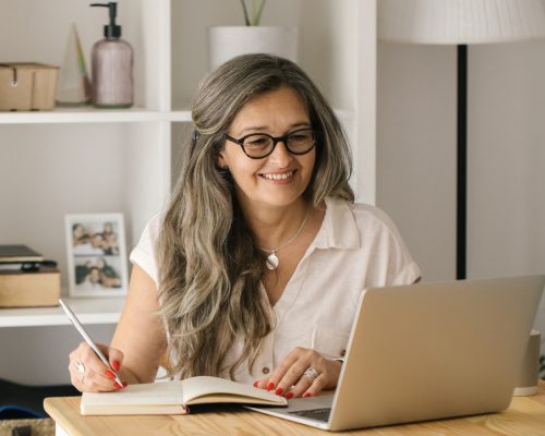 older woman working from home with a laptop and writing in a notebook