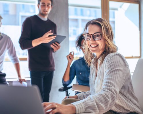 smiling person working on laptop in office while coworkers look at computer behind her